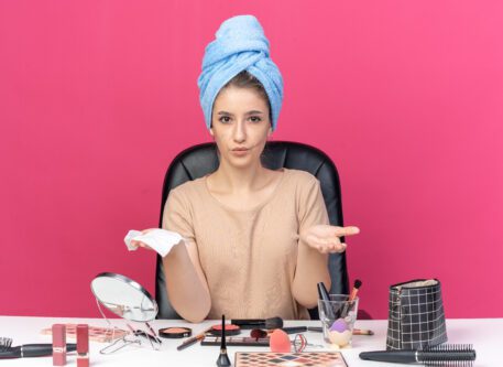 pleased young beautiful girl sits table with makeup tools wrapped hair towel holding out napkin isolated pink wall